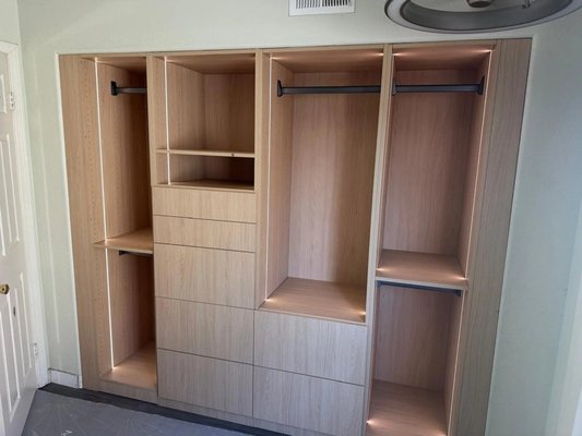 Custom Closets with Integrated Lighting - Two existing closet openings were upgraded with woodgrain storage systems, integrated lighting, drawers, open shelves, and black hanging rods. The gallery follows the original wire shelving through on-site assembly, cabinet installation, and the completed layouts. The wider closet combines multiple hanging sections with central shelving and drawers, while the smaller closet adds organized storage within its existing opening. Final photos show the finished details, including digitally styled views with clothing and jewelry. Custom Closets with Integrated Lighting - Two existing closet openings were upgraded with woodgrain storage systems, integrated lighting, drawers, open shelves, and black hanging rods. The gallery follows the original wire shelving through on-site assembly, cabinet installation, and the completed layouts. The wider closet combines multiple hanging sections with central shelving and drawers, while the smaller closet adds organized storage within its existing opening. Final photos show the finished details, including digitally styled views with clothing and jewelry.