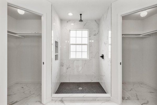Bathroom Remodel - Winnie, TX - This bathroom remodel project brings together a refined mix of modern elegance and functional design with high-end finishes and thoughtful details. The combination of marble-look tile, sleek black accents, and custom cabinetry creates a luxurious and timeless space that is both visually appealing and highly practical.

The primary bathroom features a spacious walk-in shower with frameless glass, finished with large white marble-look tiles and contrasting black hexagon floor tiles for a bold statement. A built-in shower niche with black herringbone tile adds stylish functionality. The dual vanity with shaker-style cabinetry provides ample storage, while black matte fixtures and framed mirrors bring a modern, cohesive touch to the space.

A separate makeup vanity area was incorporated, featuring a black herringbone accent wall and a round mirror, adding texture and sophistication to the space. The custom dual walk-in closets flanking the shower offer seamless storage solutions, enhancing organization and accessibility.

The secondary bathroom was designed with sleek simplicity in mind, featuring a double vanity with shaker cabinetry, quartz countertops, and matte black fixtures. The tub-shower combo is lined with the same marble-look tile, creating a cohesive aesthetic throughout the home. A horizontal transom window above the shower brings in natural light while maintaining privacy.

By using a balanced mix of textures, neutral tones, and striking black finishes, this remodel elevates both bathrooms into modern retreats. The result is a timeless, spa-like space that combines beauty and practicality in every detail.