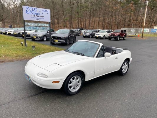1990 Mazda MX-5 Miata Convertible - This One Owner First Generation 1990 Mazda MX-5 Miata has a CARFAX Buy Back Guarantee from WS Auto Sales so you can buy with confidence that there are no undisclosed history problems. Professionally detailed and in excellent physical and mechanical condition. This 1990 Mazda MX-5 Miata is finished in Crystal White over black cloth upholstery and is powered by a 1.6-liter DOHC inline-four linked with a five-speed manual transmission and a limited-slip differential. Equipment includes 14″ Daisy alloy wheels, a black convertible soft top, air conditioning, cruise control, and an AM/FM/cassette stereo.
The MX-5 was officially introduced in February 1989 at the Chicago Auto Show and the public immediately responded with tremendous sales of new sports car. The first-generation MX-5 would go on to be the most popular of the four MX-5 generations by a wide margin, with over 228,961 units sold in the United States between 1989 and 1997. The design of the MX-5 took inspiration from the Lotus Elan, and details such as the pop-up headlights. Please call (518) 764-5929 for more information and availability. 1990 Mazda MX-5 Miata Convertible - This One Owner First Generation 1990 Mazda MX-5 Miata has a CARFAX Buy Back Guarantee from WS Auto Sales so you can buy with confidence that there are no undisclosed history problems. Professionally detailed and in excellent physical and mechanical condition. This 1990 Mazda MX-5 Miata is finished in Crystal White over black cloth upholstery and is powered by a 1.6-liter DOHC inline-four linked with a five-speed manual transmission and a limited-slip differential. Equipment includes 14″ Daisy alloy wheels, a black convertible soft top, air conditioning, cruise control, and an AM/FM/cassette stereo.
The MX-5 was officially introduced in February 1989 at the Chicago Auto Show and the public immediately responded with tremendous sales of new sports car. The first-generation MX-5 would go on to be the most popular of the four MX-5 generations by a wide margin, with over 228,961 units sold in the United States between 1989 and 1997. The design of the MX-5 took inspiration from the Lotus Elan, and details such as the pop-up headlights. Please call (518) 764-5929 for more information and availability.