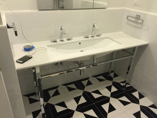 Designer Transitional Bathroom - We just wrapped up a high-end modern bathroom renovation featuring bold design choices and premium finishes throughout. The custom black and white geometric floor tile creates a dramatic, high-contrast foundation, complemented by oversized white wall tiles for a clean, seamless look that keeps the space open and bright. The floating-style vanity features dual faucets, a wide integrated trough sink, and polished chrome legs for a sleek, minimal aesthetic, paired with custom plumbing and mirrored medicine cabinets for a blend of function and style. In the shower, we installed a full spa setup with a ceiling-mounted rainhead, wall-mounted shower head, and handheld sprayer, all controlled by a sleek four-knob system for precise pressure and temperature control. The mosaic tile shower pan adds texture and slip resistance, while details like recessed lighting, integrated towel holders, and wall-mounted toilet accessories complete the custom, high-end finish.