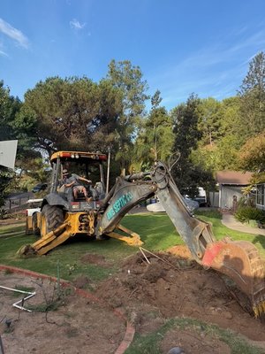200 Feet Rock & Pipe Leach Line Installation - We fixed the leach field of this beautiful homes by the installation of a 200-foot rock and pipe leach line.  We carefully excavated, as shown in the photo where our operator uses a backhoe to trench along the designated leach field area. The trench has been graded to maintain a uniform slope for proper effluent flow. Once excavated, a bed of clean drain rock is laid evenly along the trench bottom. A 4-inch perforated PVC pipe is then placed over the rock bed, with perforations facing downward to ensure even distribution. The pipe is connected and leveled using a laser or transit for accuracy. After inspection ports are installed, another 12 inches of drain rock is added to fully surround the pipe, followed by a layer of geotextile fabric to prevent soil infiltration. The trench is then backfilled with native soil and slightly mounded to allow for settling. With proper access we could complete this work in two days.