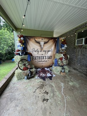 Father’s Day Backyard Western Theme - A rustic western-inspired setup featuring hay bales, wagon wheels, cowboy boots, lanterns, rope accents, and a longhorn backdrop. Finished with cow-print details and balloon garlands in earthy tones, creating the perfect country-style photo area for celebrating Dad.