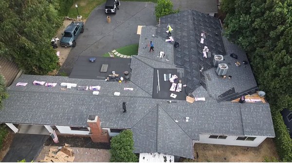 Roof Replacement in Progress – Asphalt Shingle System - This project shows an asphalt shingle roof replacement in progress across multiple roof sections. Several areas are in different stages of installation, with underlayment fully applied on some roof planes and finished shingles installed on others.

Roofing materials are staged across the surface, and crew members are actively working on shingle placement and preparation. The underlayment is evenly laid, covering large sections of the roof and providing continuous surface protection prior to shingle installation.

Existing roof features, including vents, chimneys, skylights, and HVAC units, are visible and integrated into the layout as work progresses. The project involves multiple roof angles and transitions, requiring careful alignment and coordination across intersecting roof planes.

The images reflect an organized installation process with clear staging, systematic coverage, and steady progress toward a fully completed roofing system.